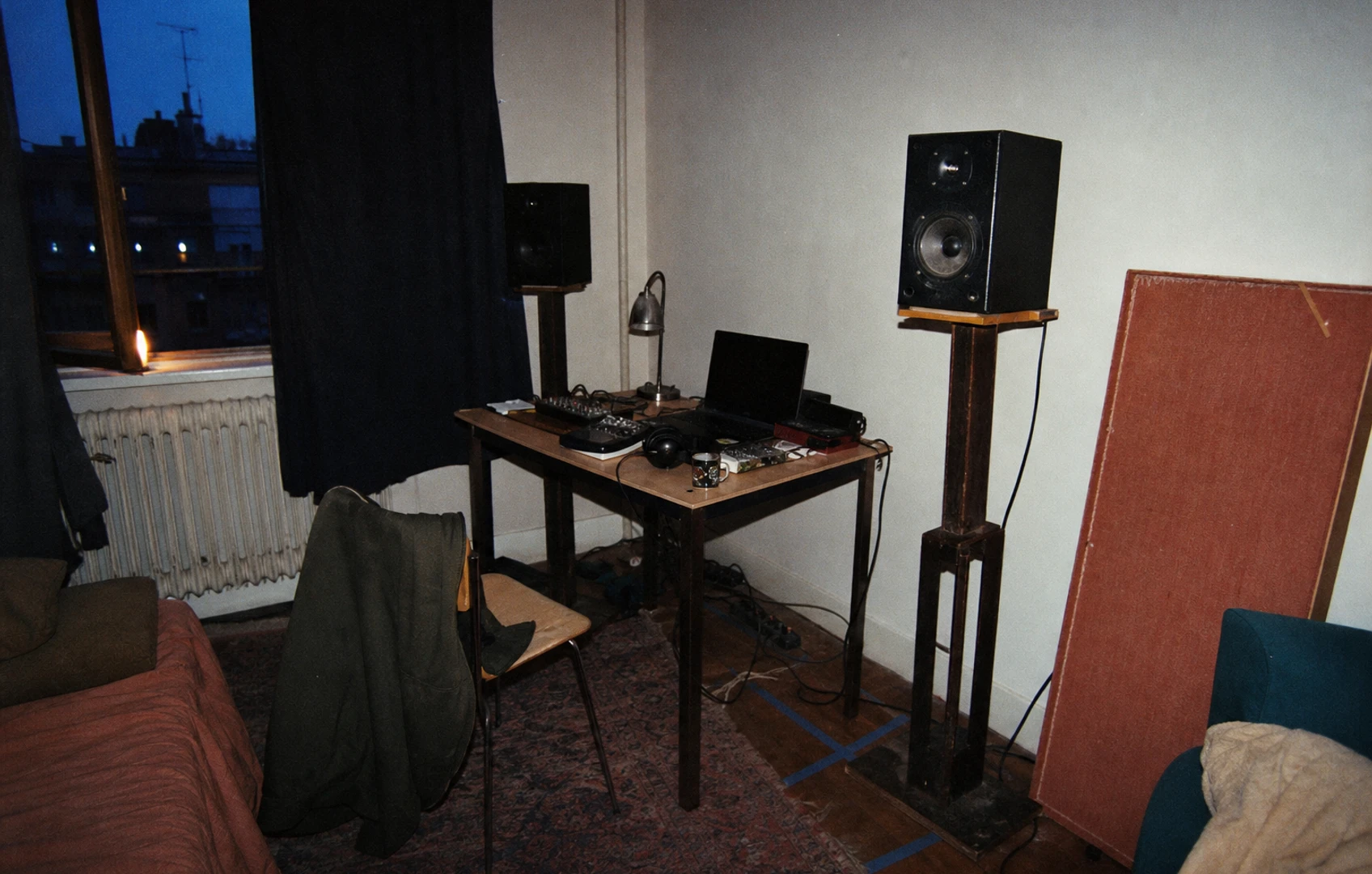 Grainy, flash-lit rented bedroom studio with mismatched monitor stands, a rumpled rug, heavy curtains, one handmade absorber, and painter’s tape marking positions on the floor.