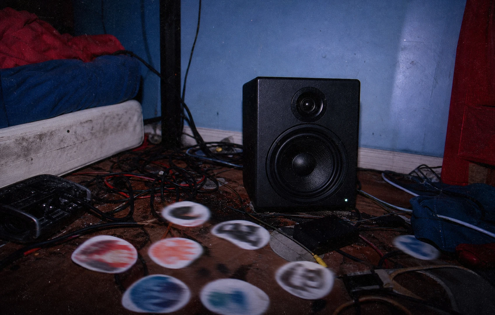 Flash-lit lo-fi bedroom studio with a monitor speaker vibrating paper circles across a cluttered desk in deep blue and red tones.