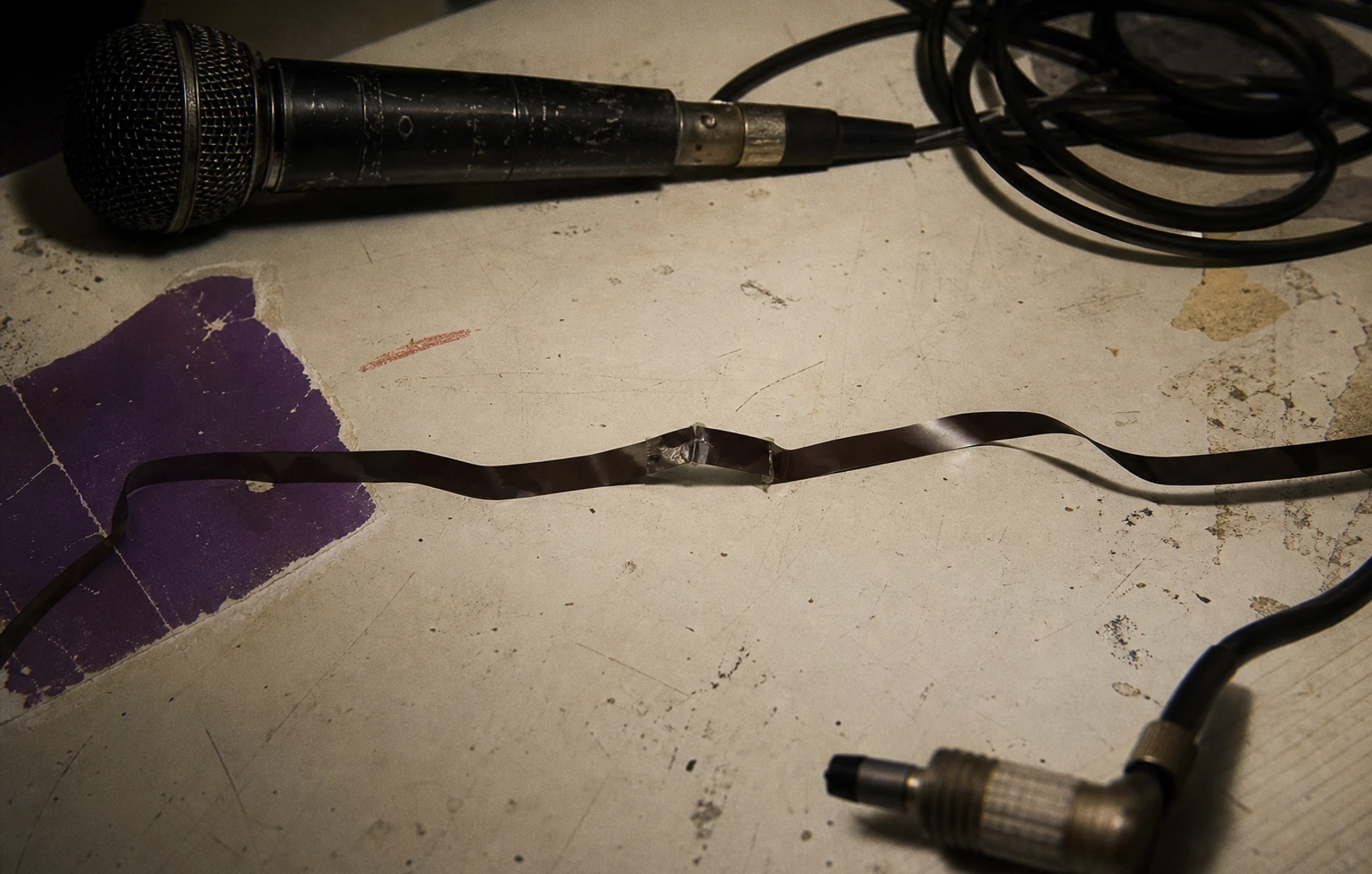 Torn-paper editorial illustration of a microphone cable, wavering tape strip, and visible hand splice in charcoal, purple, and red.