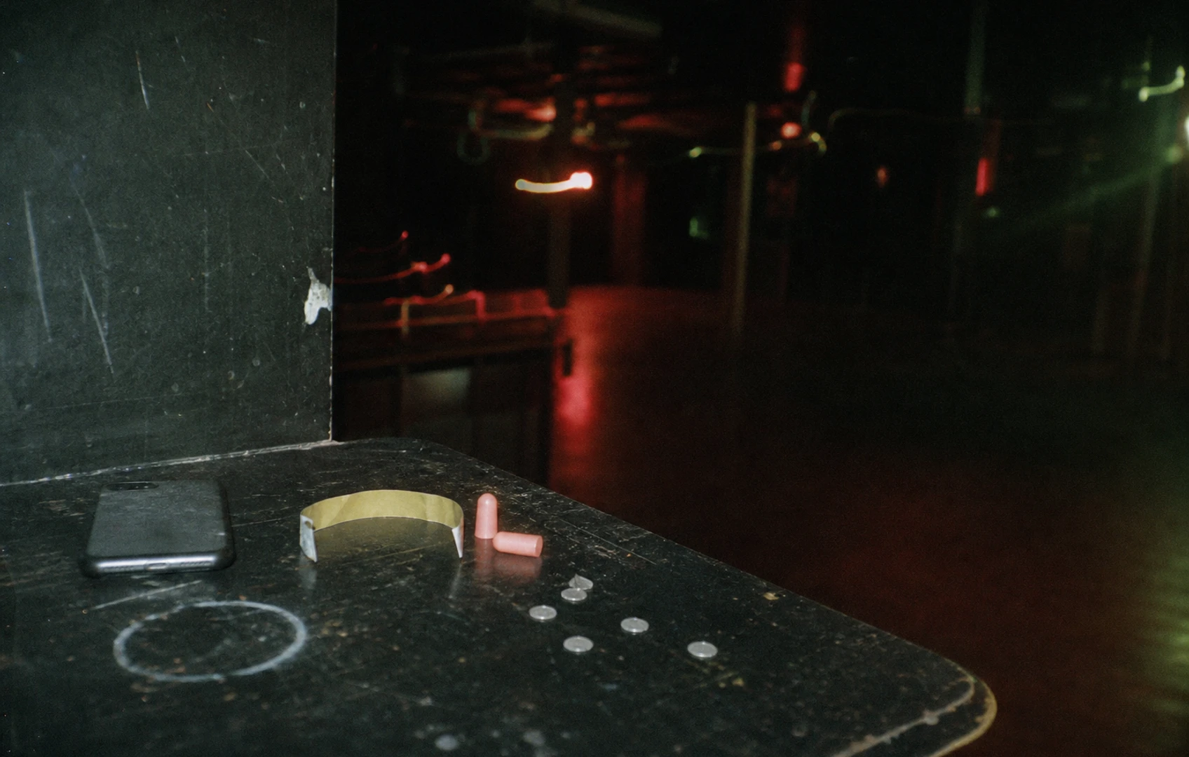 Flash-lit lo-fi still life of a face-down phone, bent wristband, foam earplugs and punched foil circles on a scratched table after a club night