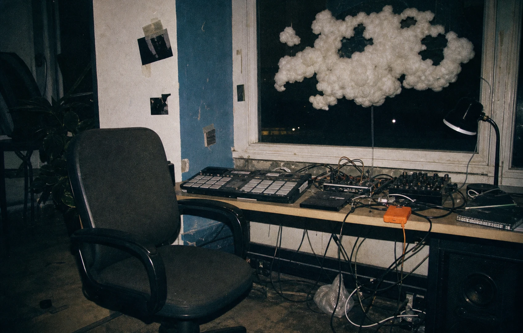 Lo-fi flash-lit studio desk with drum pads, loose cables, an external drive, and a torn paper cloud suspended above an empty chair.