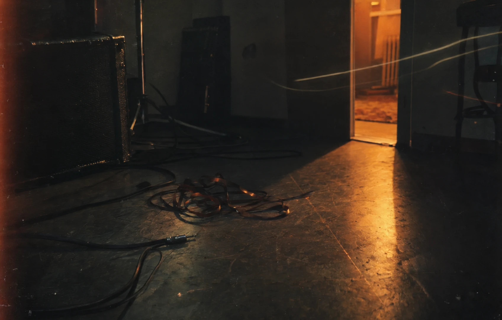 Grainy, amber-lit image of a scuffed rehearsal-room floor with a loose curl of magnetic tape beside a black instrument cable.