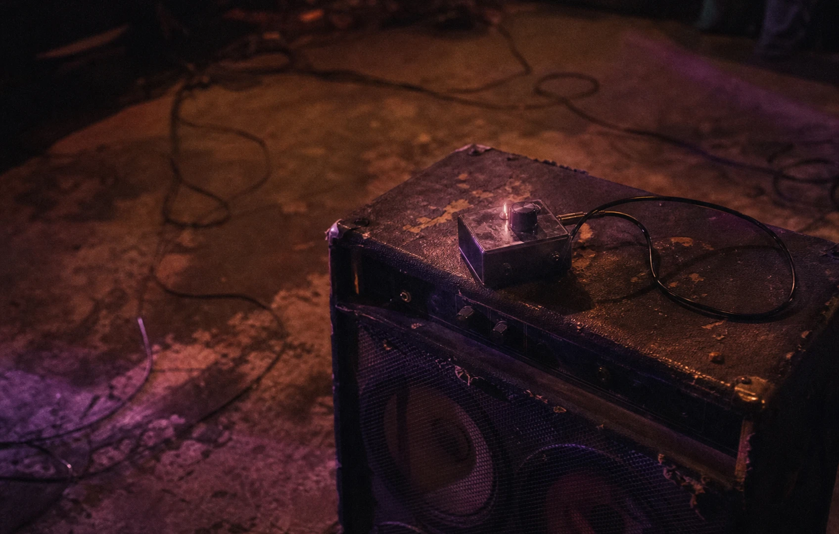 A rough editorial illustration of a small stompbox-like object glowing on a cluttered amp top, with cut-paper speaker shapes and smoky stage-light colors