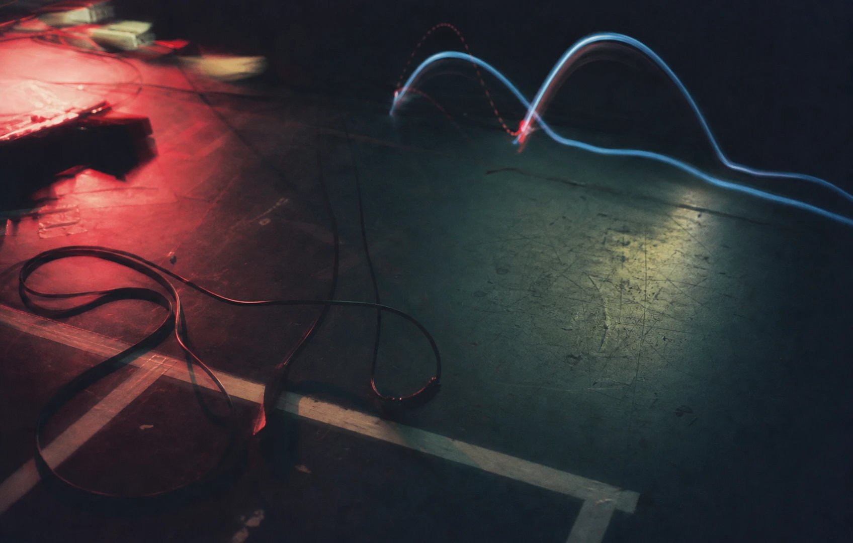 A grainy editorial collage of looping instrument cables thinning into abstract radio waves over a dark stage floor marked with tape lines and pedal shadows.
